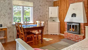 Ein gemütliches Zimmer mit einem Holzesstisch, Stühlen, einem weißen Schrank, einem Kamin und einem Fenster mit Blick auf Grünflächen.