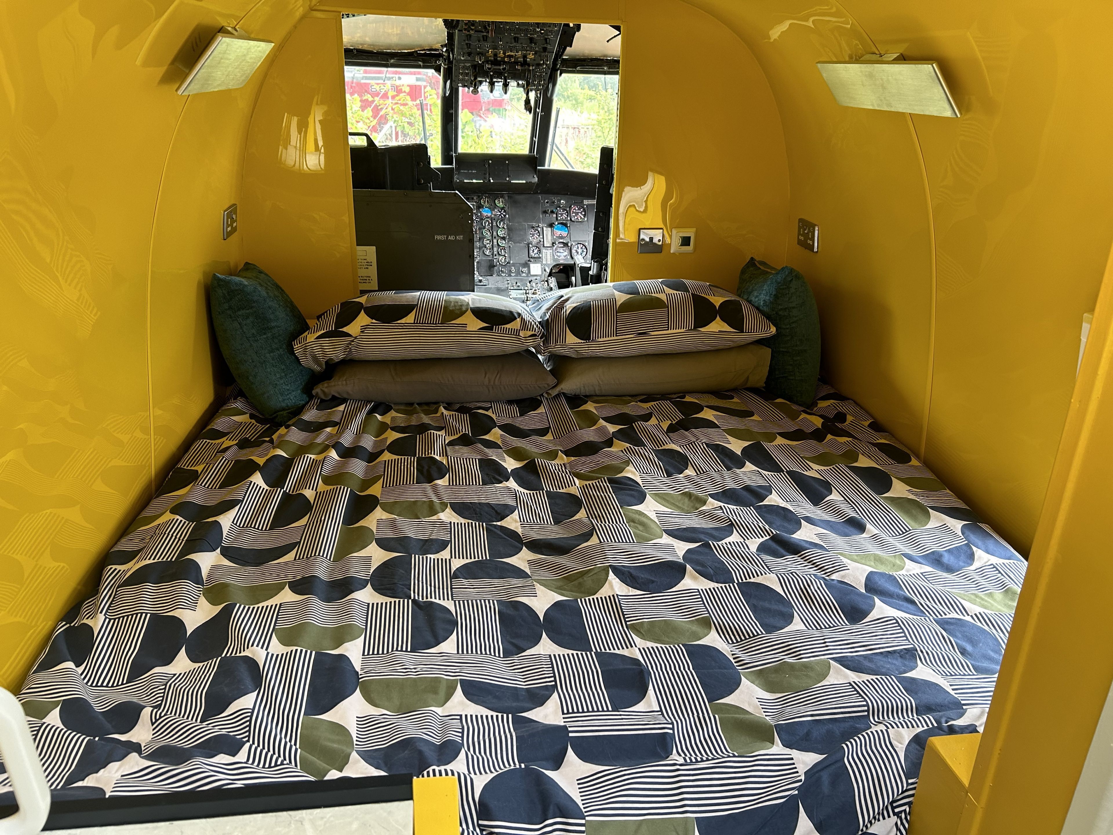 Un intérieur compact aux couleurs chaudes, avec un lit agrémenté d’une couvre-lit à motifs géométriques et de coussins assortis.