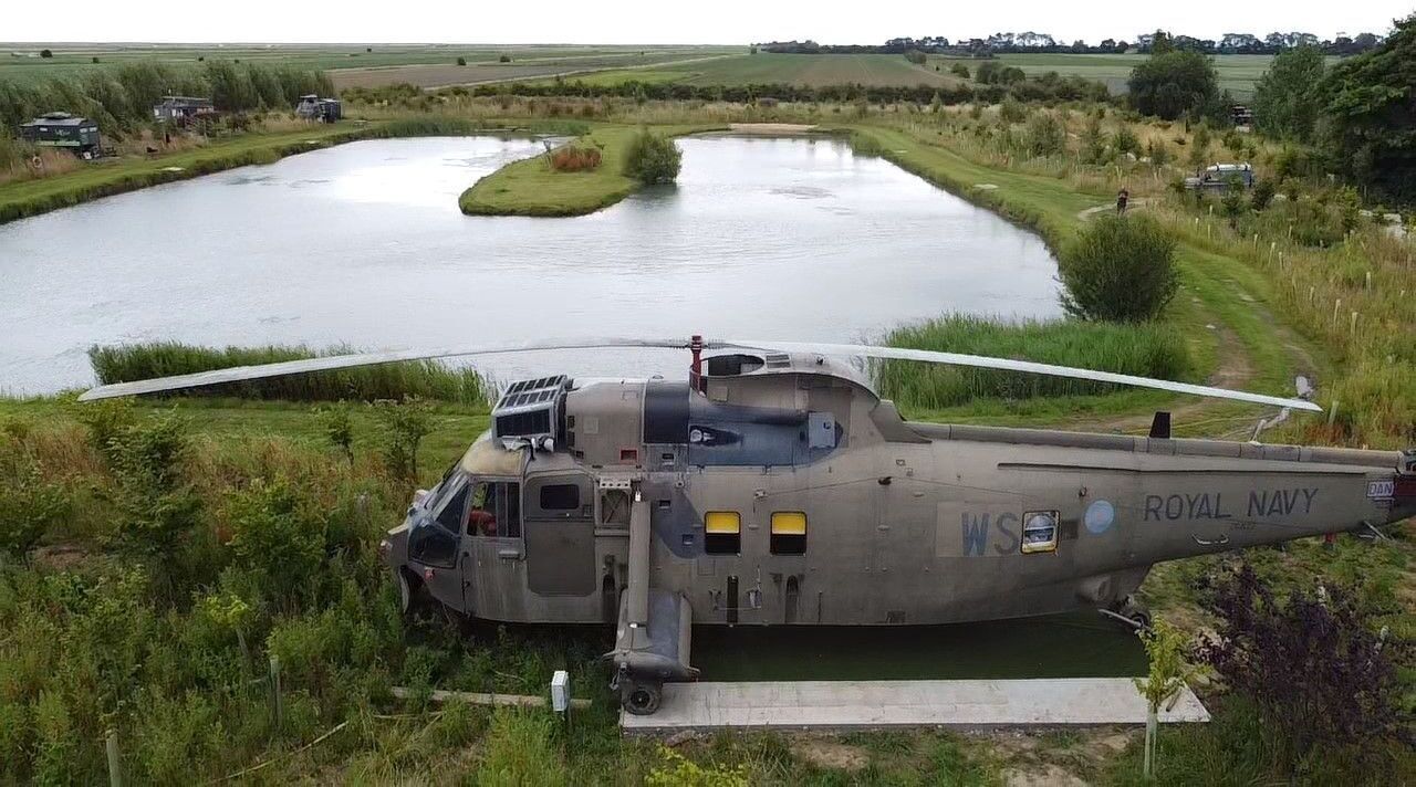 A military helicopter with the markings "Royal Navy" and "WS" parked near a body of water, surrounded by greenery and a small building.