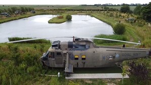 Un hélicoptère militaire portant les inscriptions « Royal Navy » et « WS » est stationné près d’une étendue d’eau, entouré de verdure et d’un petit bâtiment.