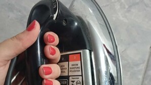 Une main tenant un fer à repasser noir, sur lequel est apposée une étiquette indiquant les réglages à utiliser pour différents types de tissus.
