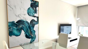 A modern living room with a glass dining table, white chairs, a large abstract painting, a flat-screen TV, and a window with a view. - Azores Paim Apartment T2 21A (Ponta Delgada)