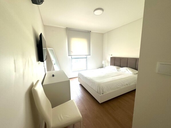 A modern hotel room with a bed, a desk, a chair, a TV, and a window with blinds.