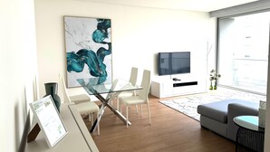 A modern living room with a glass dining table, white chairs, a grey sofa, a flat-screen TV, and a large abstract painting on the wall. - Azores Paim Apartment T2 21A (Ponta Delgada)