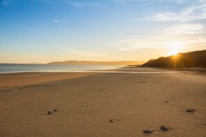 A wide sandy beach with gentle waves, a clear sky, and a distant sun setting or rising. - 5 Parc Delfryn - sleeps 7 guests in 3 bedrooms (Benllech)