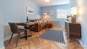 Un salon moderne avec un parquet en bois disposé en chevrons, un canapé en cuir marron, une table à manger avec des chaises, un tapis à motifs et une lampe.