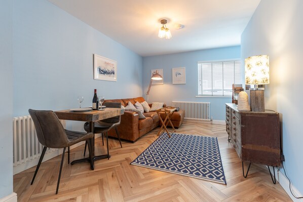Een moderne woonkamer met een houten visgraatvloer, een bruine leren bank, een eettafel met stoelen, een getextureerd vloerkleed en een lamp.