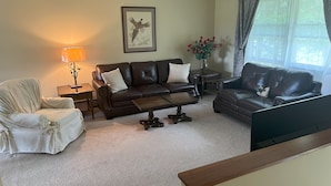 Un salon avec un canapé en cuir marron, un fauteuil blanc, une table basse, une lampe, un tableau encadré au mur et une fenêtre avec des rideaux.