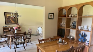 Un coin repas avec une table et des chaises en bois, un lustre, une étagère remplie d’assiettes et un tableau accroché au mur.