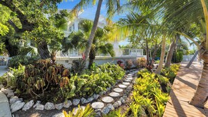 A tropical garden pathway with a wooden deck, surrounded by various plants and palm trees. - Reel Joy | Florida Keys Waterfront Home + Pool & Private Dock (Marathon)