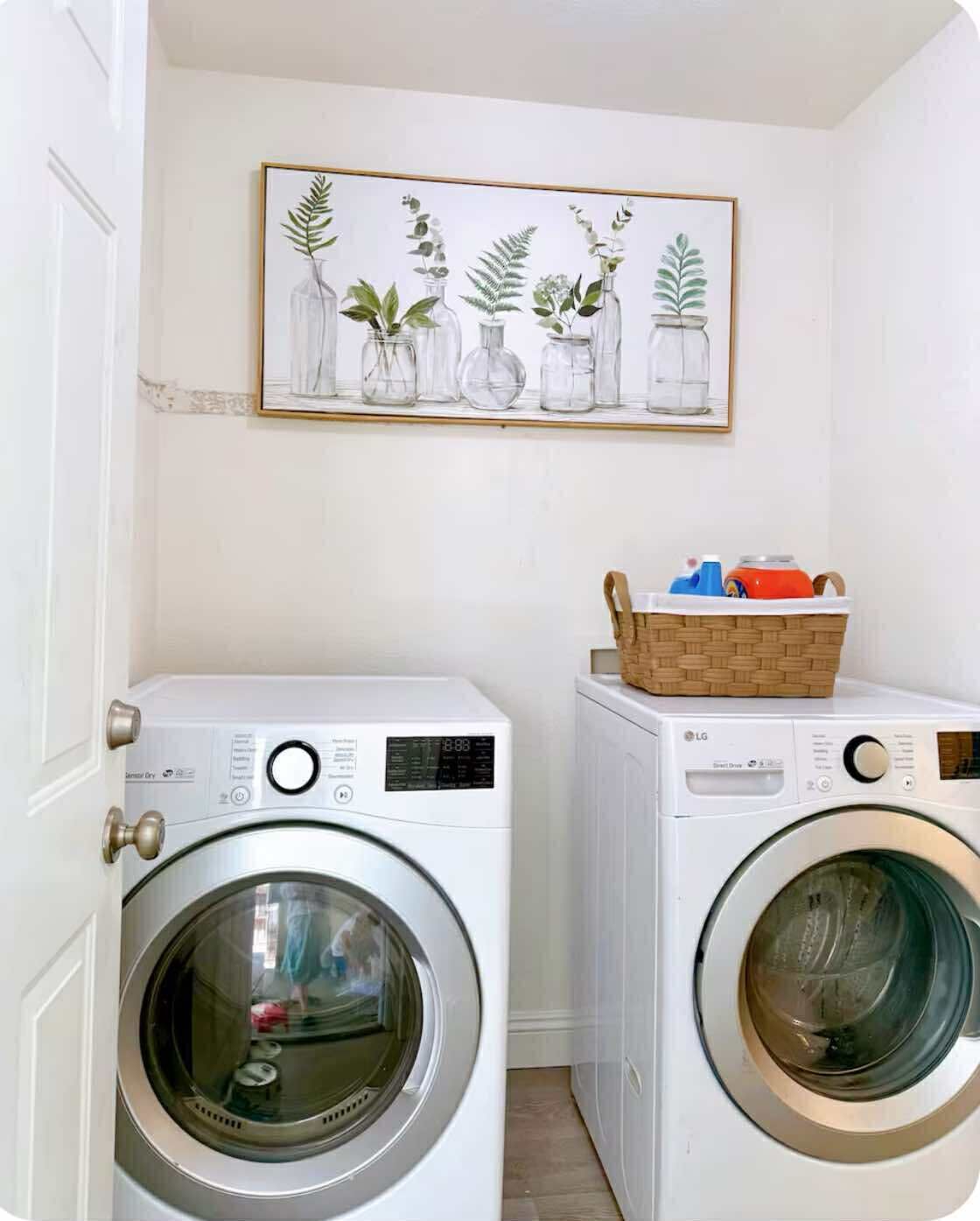 Ein Waschraum mit zwei Waschmaschinen mit Frontlader, einem Korbwäschesammler für Reinigungsmittel und einem gerahmten Pflanzenmotiv an der Wand.
