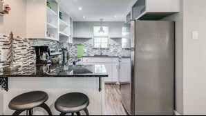 A modern kitchen with white cabinetry, a stainless steel refrigerator, a black countertop, and two bar stools.