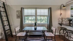 A dining area with a table, chairs, a ladder, a window with curtains, and a kitchen area with stools.