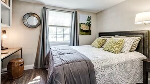 A bedroom with a bed, a desk, a chair, a lamp, a window with curtains, and a painting on the wall.