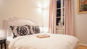 Una cama bien hecha con una bandeja de tazas y platillos, una mesita de noche y una ventana con cortinas.