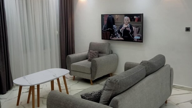 A modern living room with a grey sofa, a grey armchair, and a small white table. A flat-screen TV is mounted on the wall, and there are two wall-mounted lights.