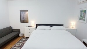 A bedroom with a bed, a grey sofa, two bedside tables with lamps, a small trash bin, and a rug. - The Williams Center (New London)