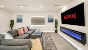 Ein modernes Wohnzimmer mit grauem Sofa, einem Flachbildfernseher an der Wand und einem Kamin mit blau beleuchtetem Bildschirm.