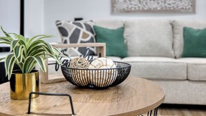 A round wooden table with a black wire basket containing decorative items, a gold-colored vase with a plant, and a framed picture on a light-colored sofa with green cushions. - Military in Style/Movie Room /Game Room Minutes from Ft. Benning & Downtown area (Columbus)