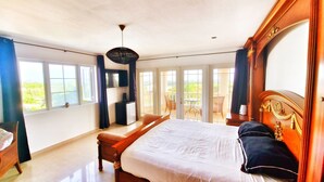 Une chambre spacieuse avec un grand lit, une tête de lit en bois, un bureau et une chaise. Elle dispose d’un balcon avec une table et des chaises, et offre une vue sur la verdure environnante.