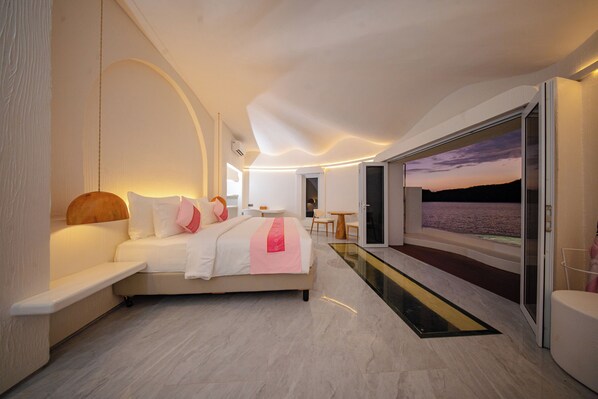 A modern bedroom with a large bed, a bedside table, a chair, and a view of the sea through a glass door. - 69 Resort & Beach Club by Loccal Collection, Labuan Bajo (Labuan Bajo)