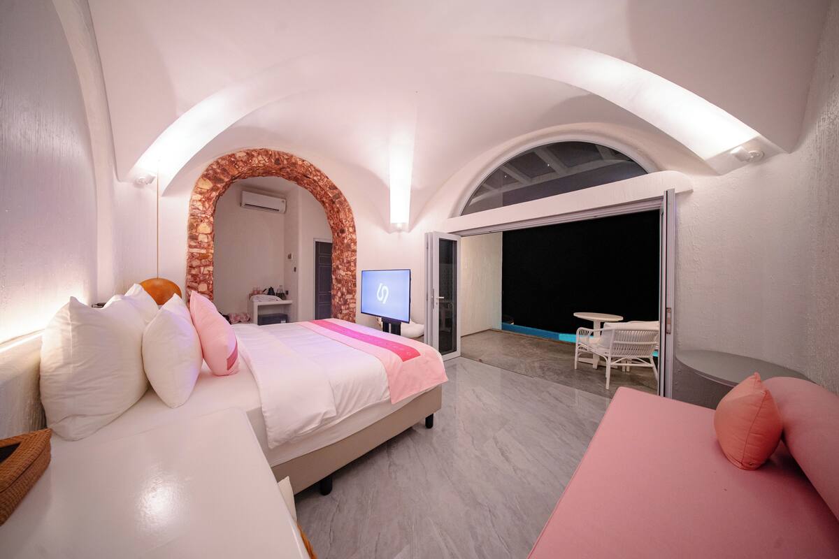 A bedroom with a bed, a TV, a balcony with outdoor furniture, and a stone archway.