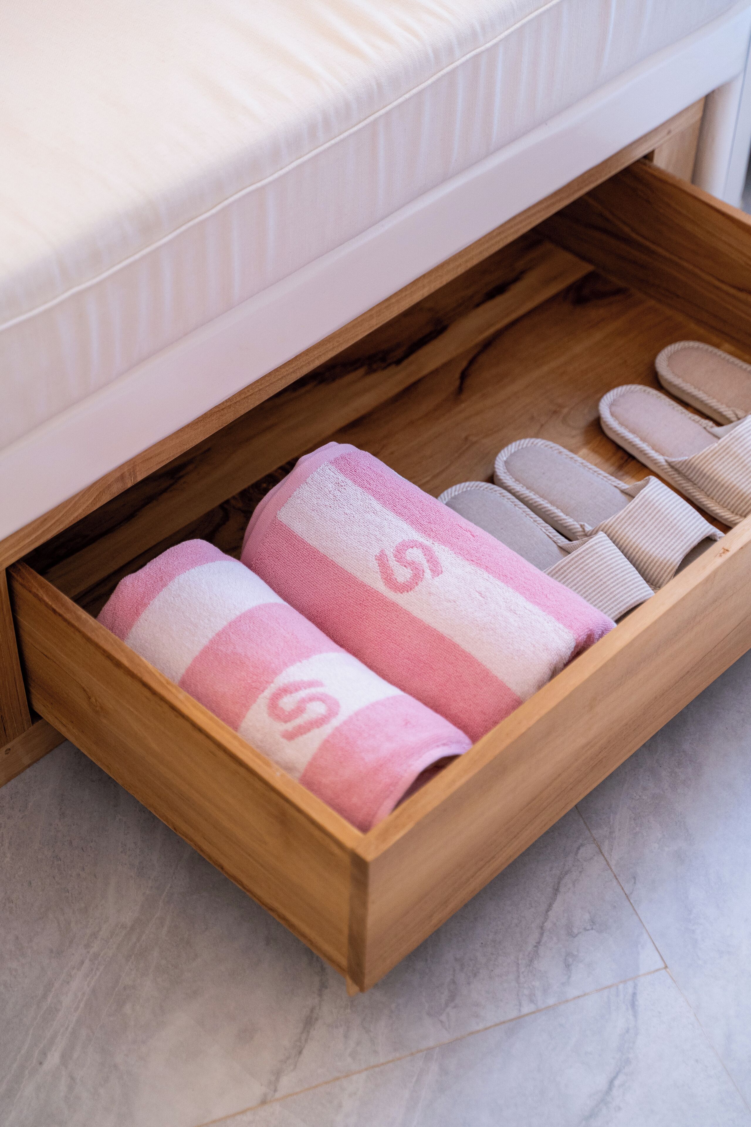 A wooden drawer under a bed containing pink and white striped towels and slippers.
