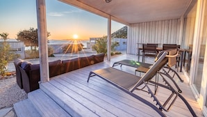 A wooden deck with a lounge chair and a table, overlooking a body of water and a sunset.