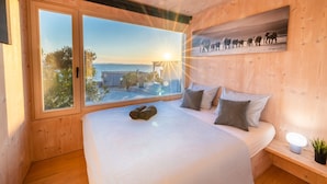 A bedroom with a large window, wooden walls, a bed with pillows, and a small bedside table with a plant.