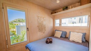 Dormitorio de cabaña de madera con cama, dos almohadas, decoración de macramé colgante y ventana con vistas a follaje.