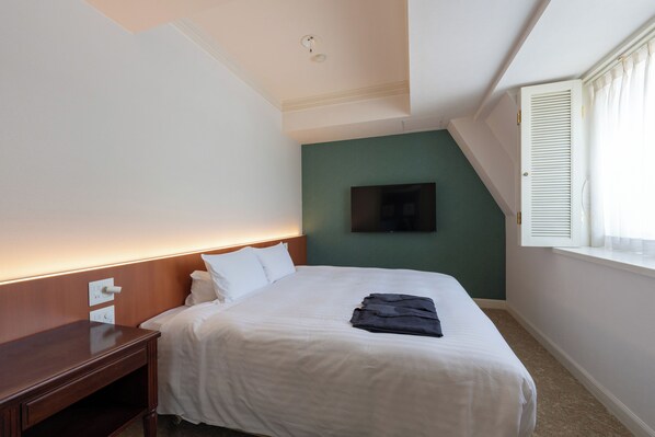 A hotel room with a neatly made bed, a wooden nightstand, a flat-screen TV mounted on the wall, and a window with white shutters.
