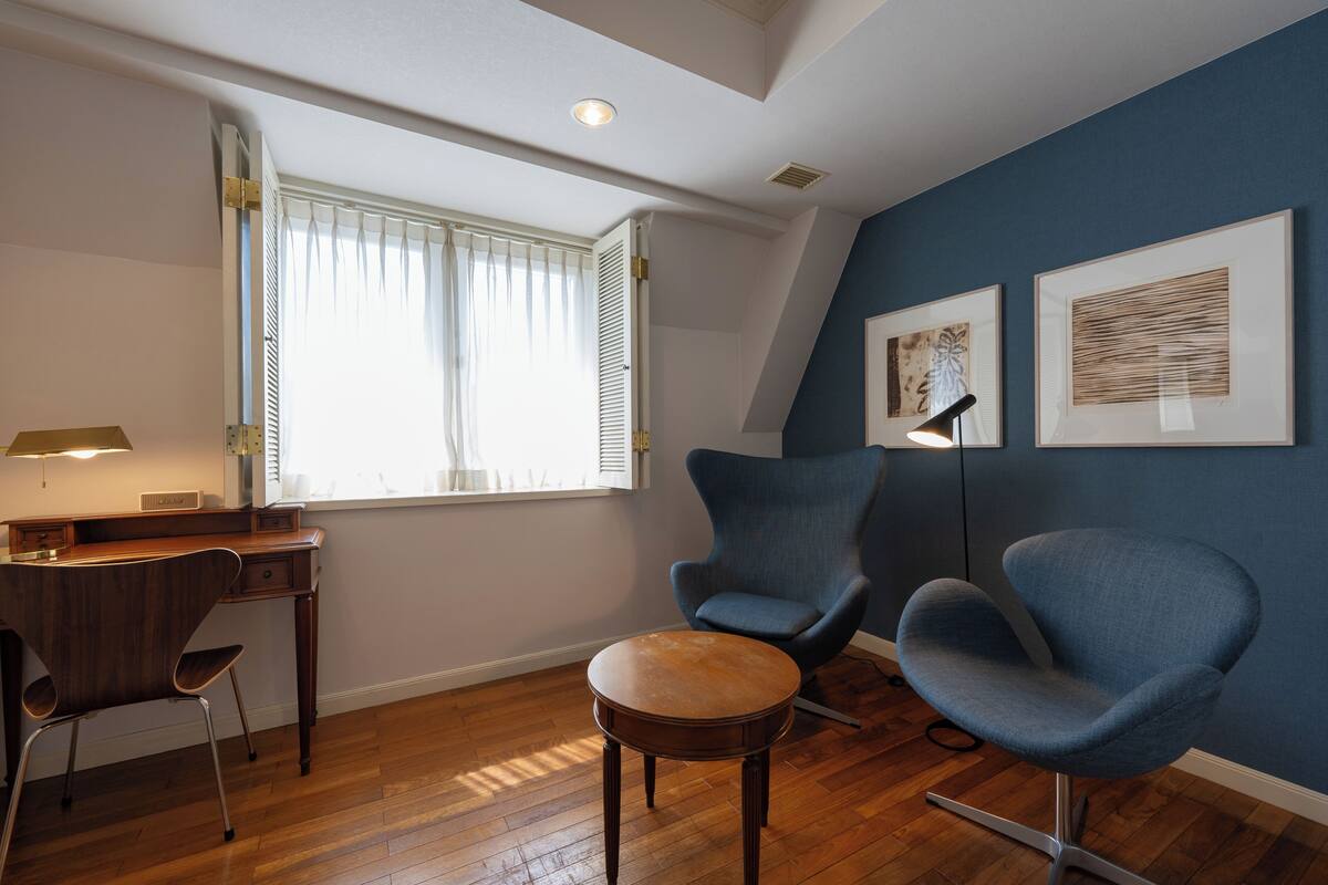 Ein Zimmer mit einem Holztisch, zwei blauen Stühlen, einem kleinen runden Tisch, einem Fenster mit weißen Rollläden und zwei gerahmten Kunstwerken an der Wand.
