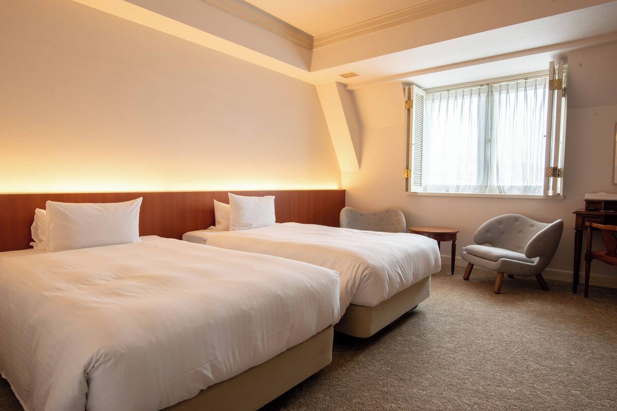 Ein Hotelzimmer mit zwei Betten, einem kleinen Tisch, einem Stuhl und einem Fenster mit Vorhängen.
