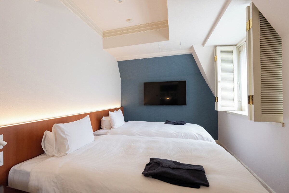 Una habitación de hotel con cama, televisor de pantalla plana montado en la pared y ventana con contraventanas.