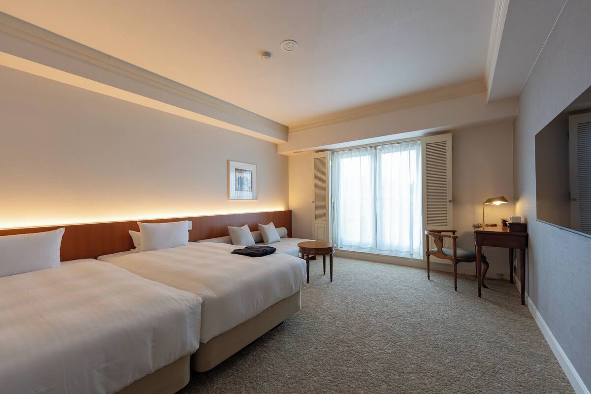 Ein Hotelzimmer mit zwei Betten, einem kleinen Tisch, einem Sessel und einem Schreibtisch.