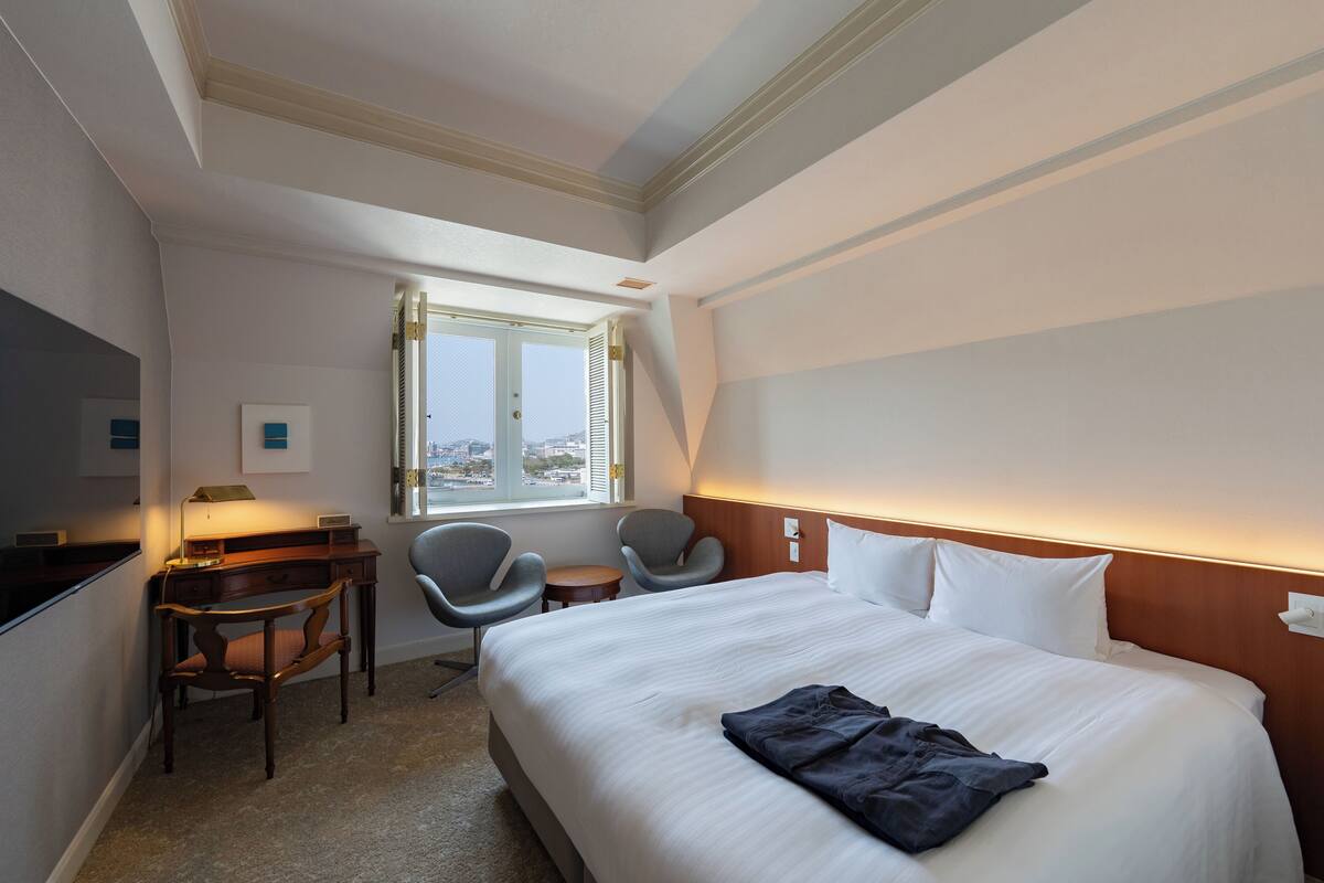 Ein Hotelzimmer mit Bett, Schreibtisch, Stühlen und einem Fenster mit Blick auf die Stadt.