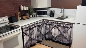 Una cucina con piano cottura bianco, forno a microonde e frigorifero. Si nota un mobiletto in legno e una parete in mattoni.