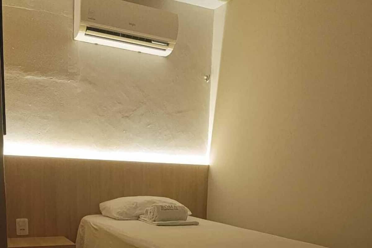 A small, neatly made bed in a corner room with a built-in air conditioning unit above it.