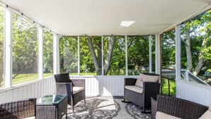 Un espacio al aire libre cubierto con mobiliario de mimbre y una mesa de vidrio, rodeado por barandillas blancas y grandes ventanales.