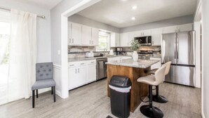 Una cocina moderna con gabinetes blancos, una isla central, electrodomésticos de acero inoxidable y una silla gris.