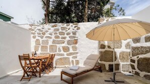Un mur de pierres, une table en bois, des chaises, un fauteuil et un grand parasol, le tout dans un décor extérieur.