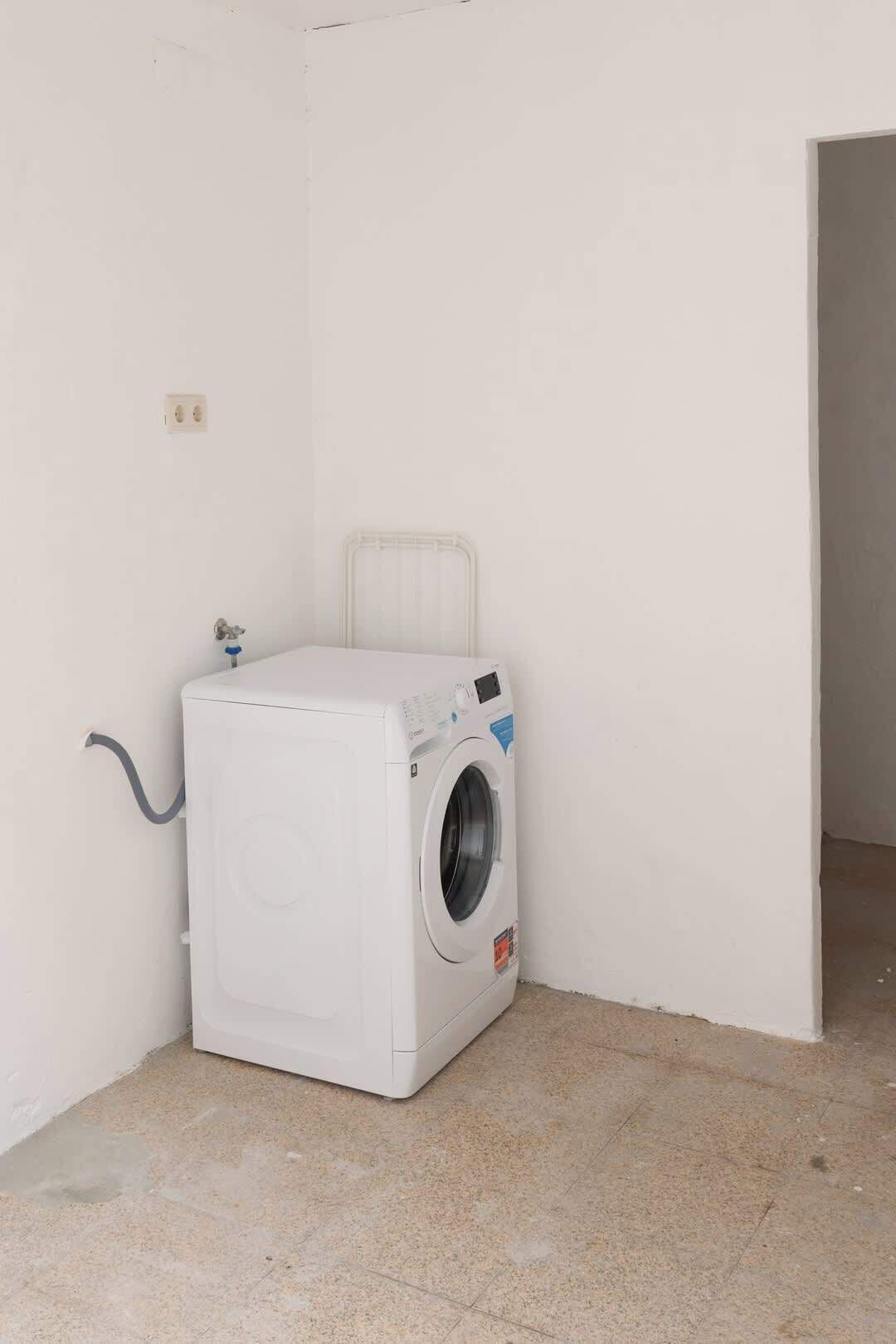 A compact, white washing machine in a corner of a room with tiled flooring and plain walls.