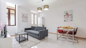 Una sala moderna con un sofá gris, una mesa de centro de vidrio y una zona de comedor con una mesa blanca y sillas rojas. Hay un cuadro en la pared y una ventana con cortinas blancas.
