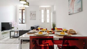 Un comedor moderno con una mesa blanca preparada para el desayuno, sillas rojas y un televisor al fondo.