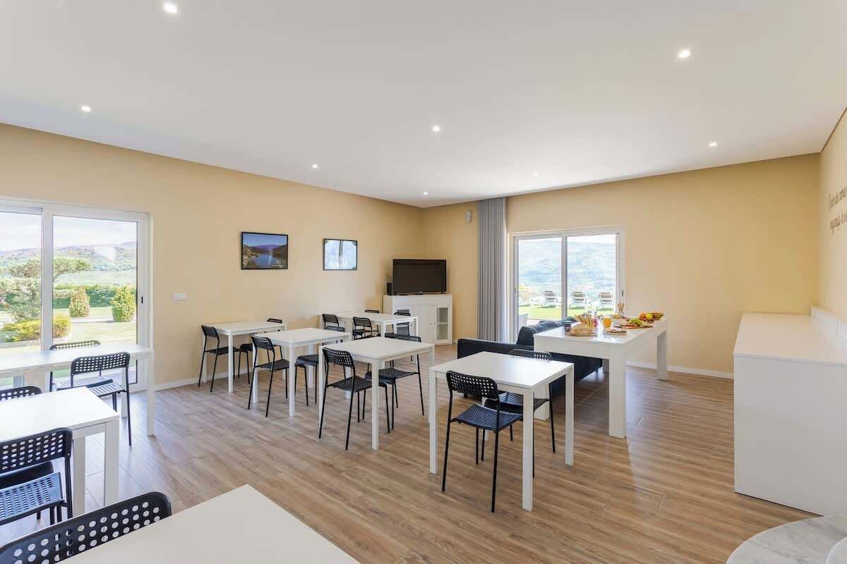 Uma área de jantar moderna com várias mesas e cadeiras, uma televisão de tela plana e grandes janelas com vista para o exterior.