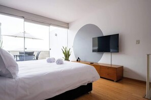 A modern hotel room with a large bed, a flat-screen TV mounted on the wall, a wooden dresser, and a balcony with a table and chairs.