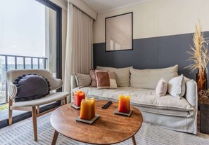 A modern hotel room with a sofa, chair, round coffee table, and a large window with a view.