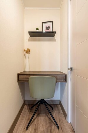 A corner desk with a chair, a small shelf with a picture, and a vase with dried plants.