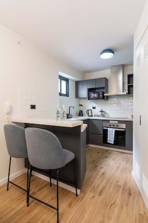 Una cocina compacta con una isla central, dos taburetes de bar, microondas, hervidor y horno empotrado.