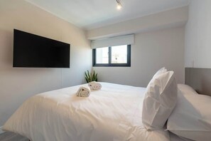 Un dormitorio con una cama, un televisor de pantalla plana montado en la pared, una ventana con persianas y una planta.